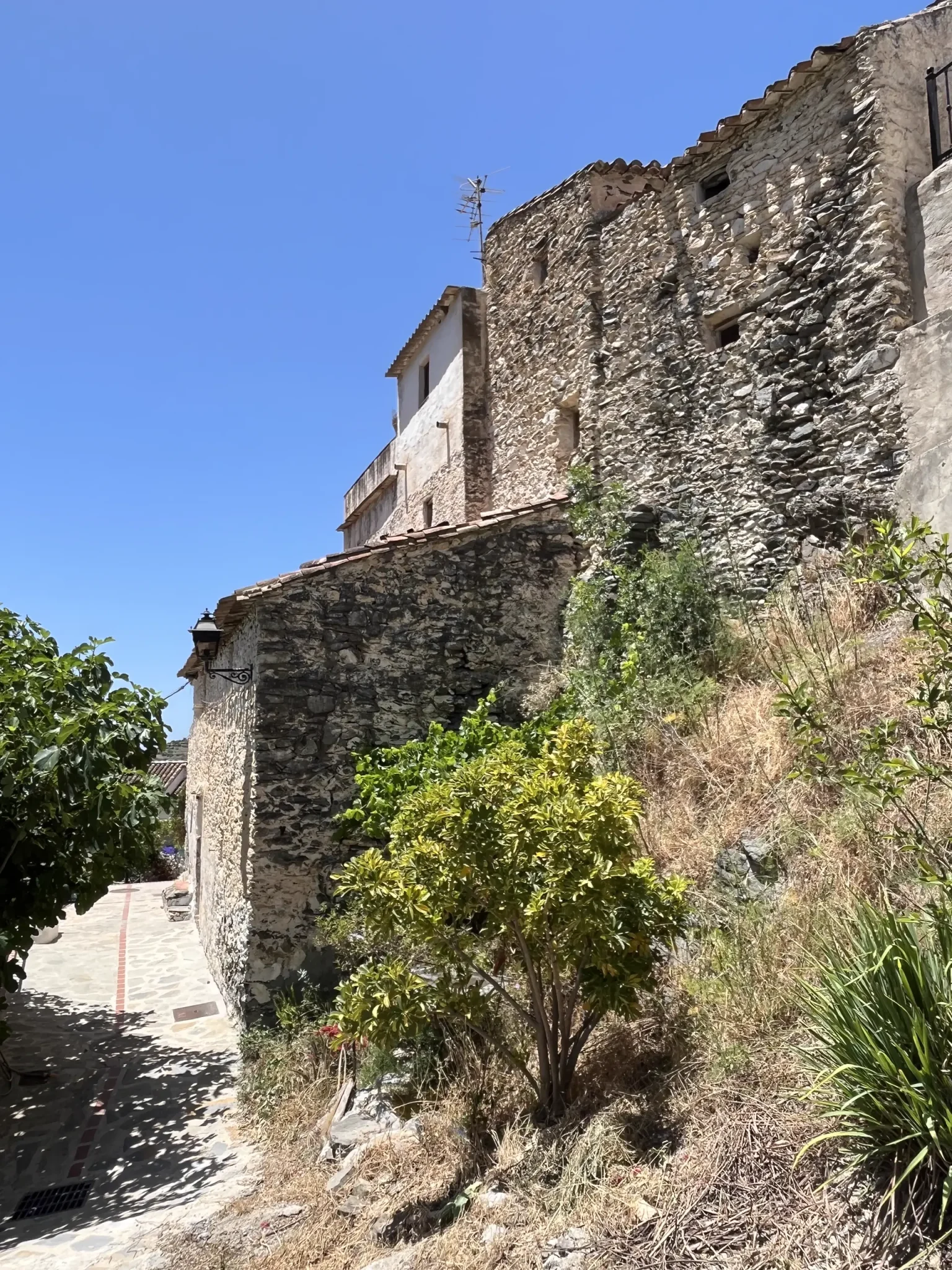 Casa con muros de mampostería de piedra sobre un talud con vegetación, junto a una calle empedrada de pueblo